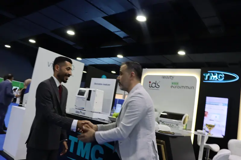 Two men shaking hands and smiling in front of the TMC counter and the Revvity IDS and EUROIMMUN wall